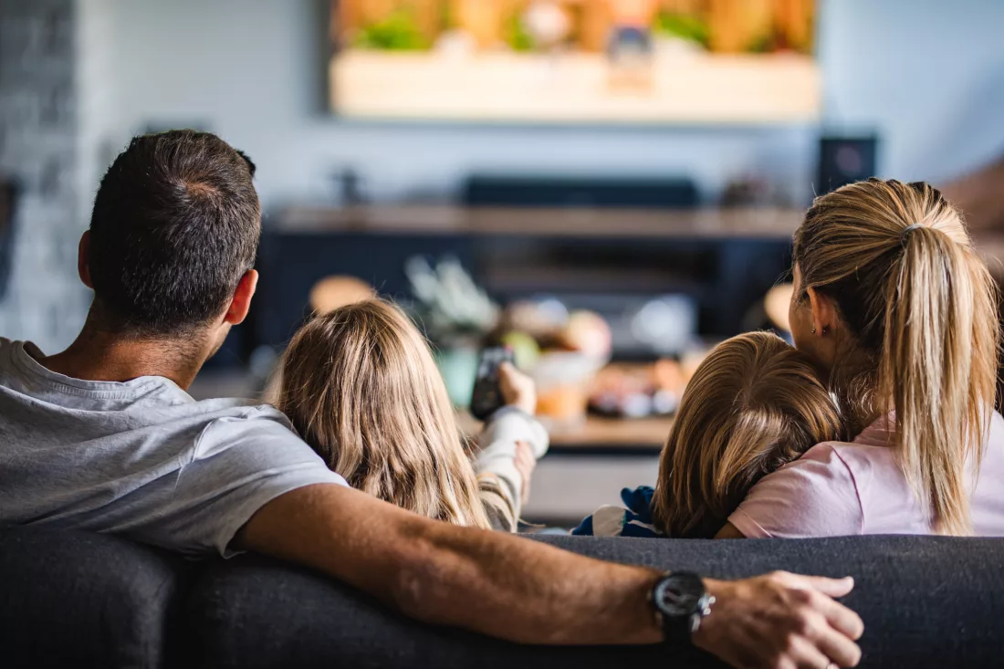 Na imagem, uma família sentada no sofá assistindo aula na TV