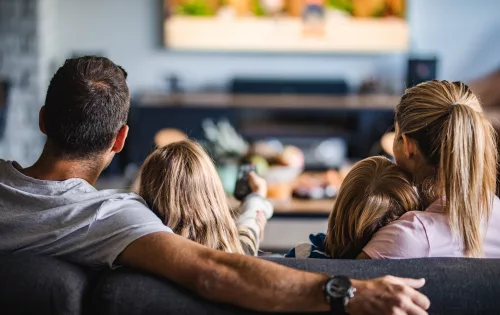 Na imagem, uma família sentada no sofá assistindo aula na TV