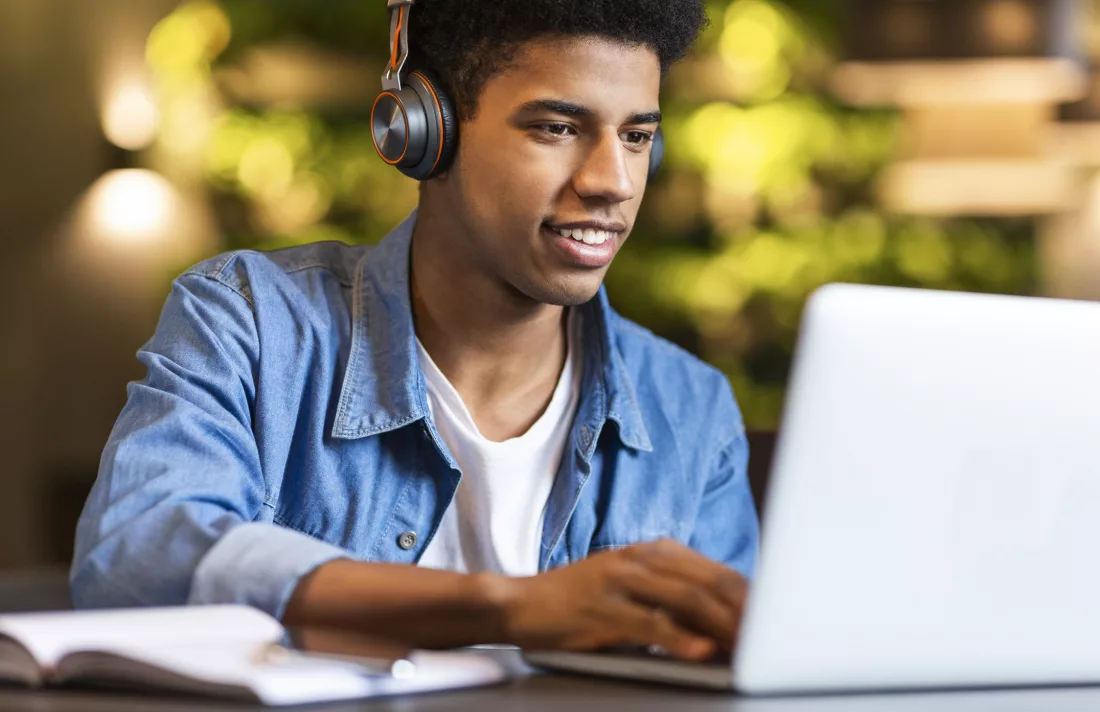 Na imagem, um jovem sentado na frente do computador, com fones no ouvido e acompanhando cursos gratuitos