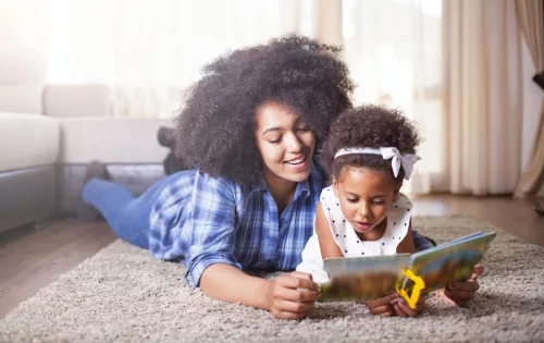 Na imagem, uma mãe com a sua filha estão deitadas no chão com um livro de fábulas nas mãos