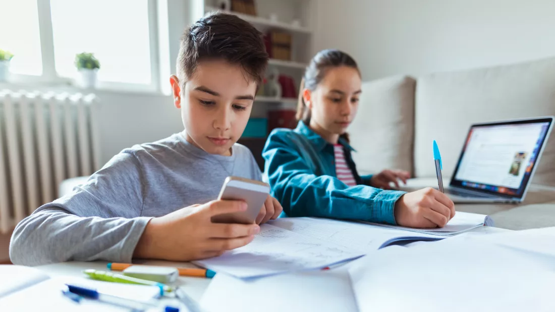 Na imagem aparece um menino estudando com celular na mão, ao lado de uma menina que se apoia nos cadernos