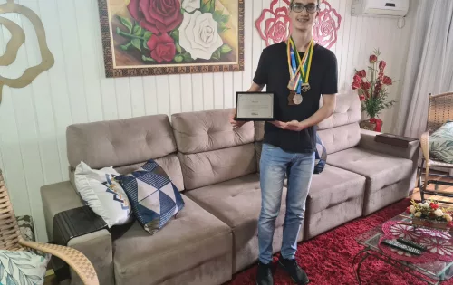 Menino de 16 anos, na sala de casa, segurando certificado e várias medalhas que ganhou em olimpíadas científicas