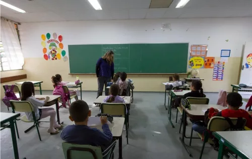 Na foto, alunos estão em uma sala de aula acompanhados de uma professora