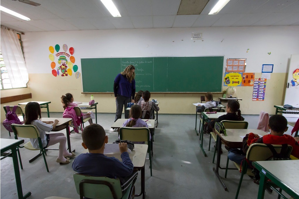 Na foto, alunos estão em uma sala de aula acompanhados de uma professora