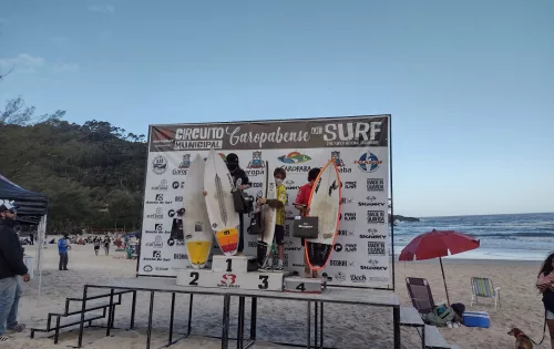 Surfistas em cima de um pódio, recebendo a premiação de um campeonato de surfe na Praia da Ferrugem em Garopaba