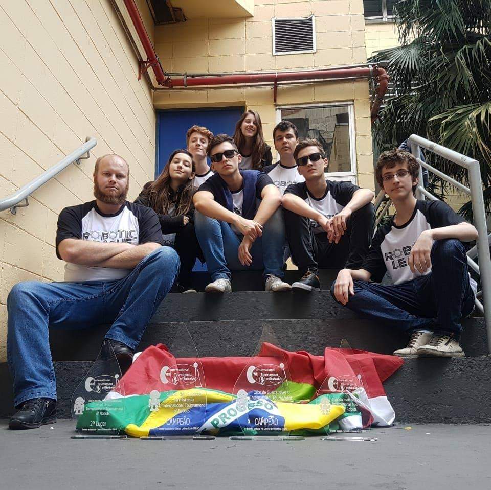 Alunos da oficina de robótica e astronáutica do professor Carlos Rutz sentados na escada com a bandeira do Brasil e do estado de Santa Catarina e troféu internacional do International Tournment of Robots em 2018