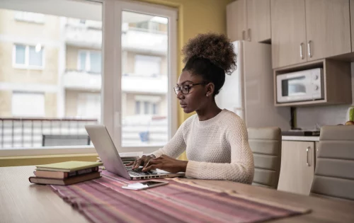 Mulher negra sentada na mesa da cozinha de casa, em frente ao computador, fazendo cursos online e gratuitos de capacitação