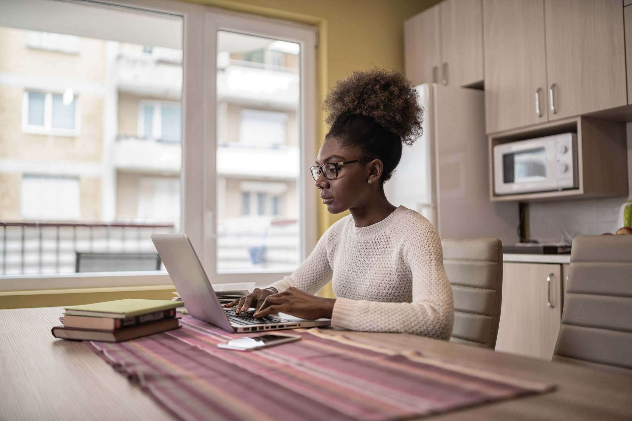 Mulher negra sentada na mesa da cozinha de casa, em frente ao computador, fazendo cursos online e gratuitos de capacitação