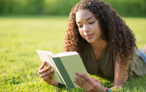 Menina adolescente deitada em um gramado com um livro aberto nas mãos