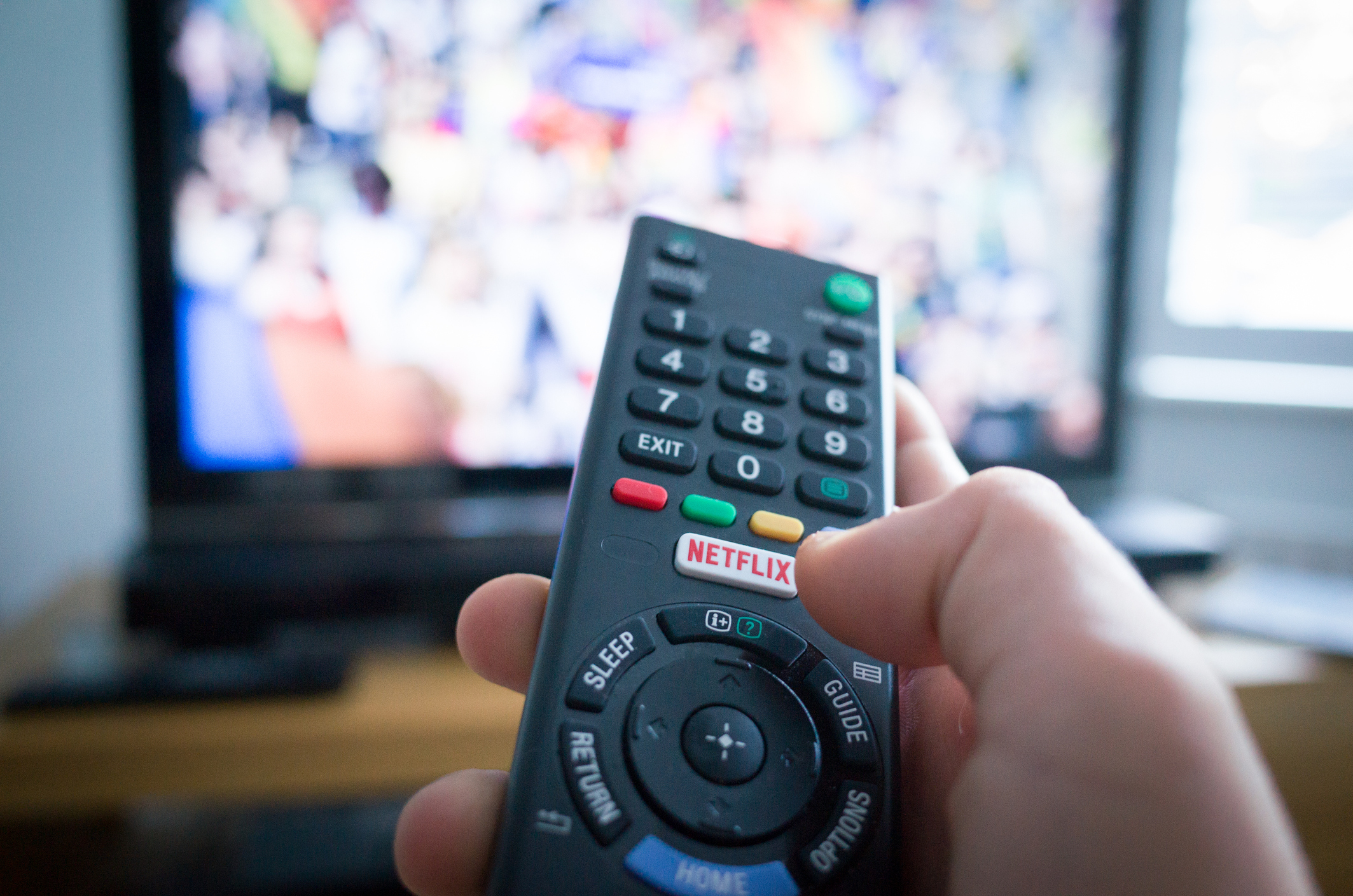 Mão segurando um controle remoto apontado para uma TV, escolhendo séries para assistir na Netflix durante o recesso escolar 