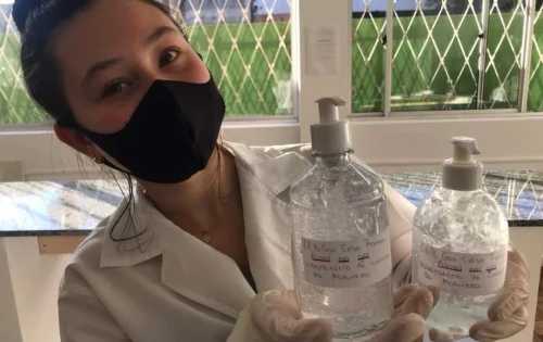 Estudante Nely Ribeiro Morales, 16, da segunda série do Novo Ensino Médio da EEB Celso Ramos, de Joaçaba, vestindo jaleco, luvas e máscara, e segurando dois recipientes com álcool em gel produzidos pela turma no laboratório de ciências da natureza da instituição de ensino