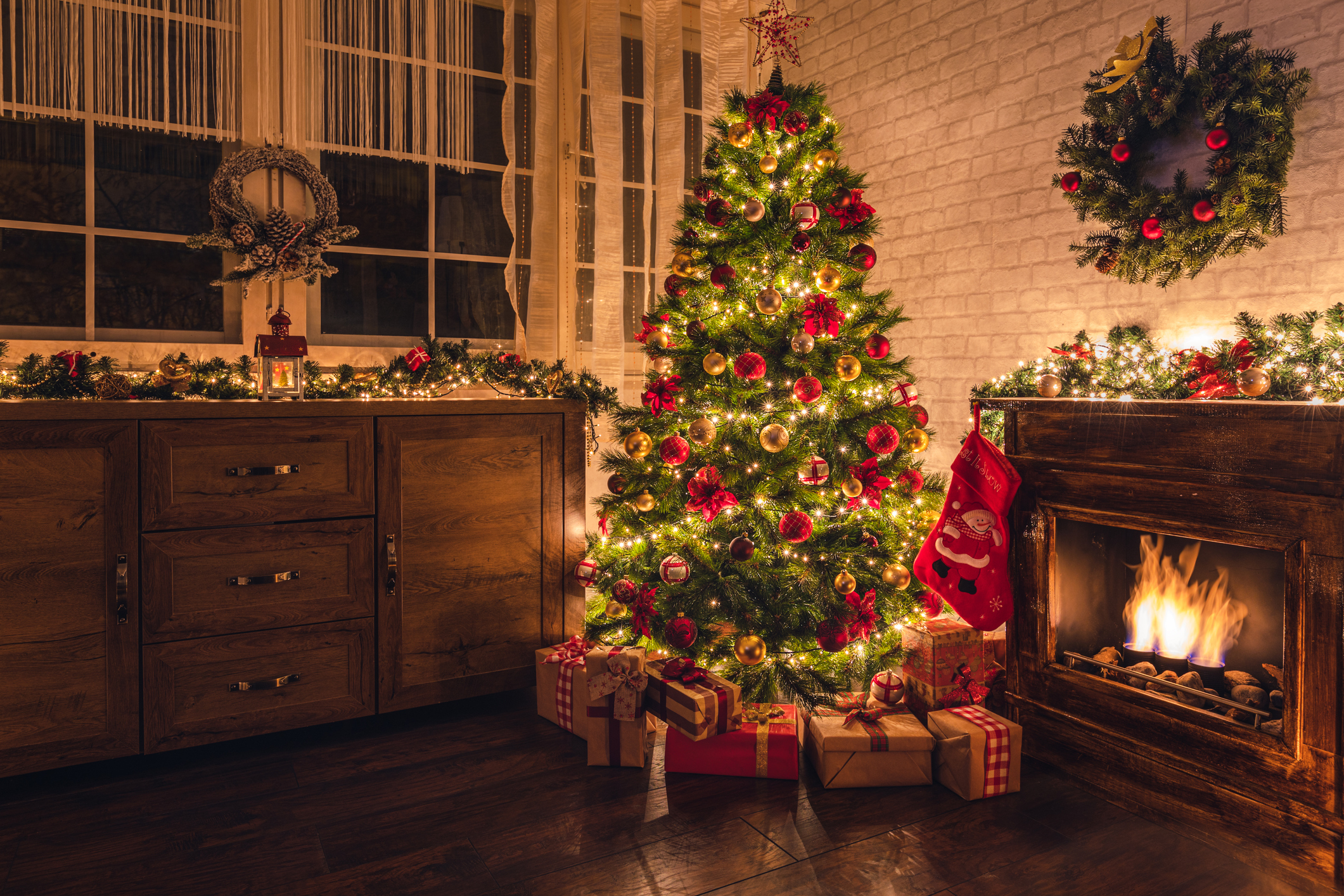Imagem mostra uma árvore de Natal, decorada com luzes, bolas e outros enfeites, na sala de uma casa com lareira e outros enfeites de Natal. Embaixo da árvore estão os presentes de Natal.