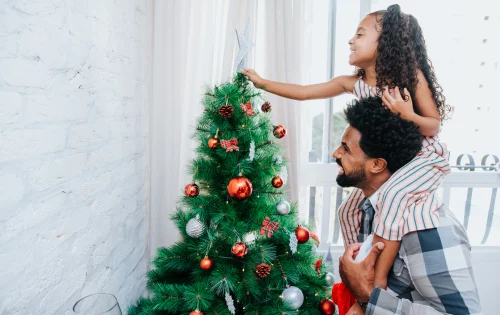 Imagem mostra uma criança nos ombros do pai colocando uma estrela no topo de uma árvore de natal decorada com bolas de diversas cores