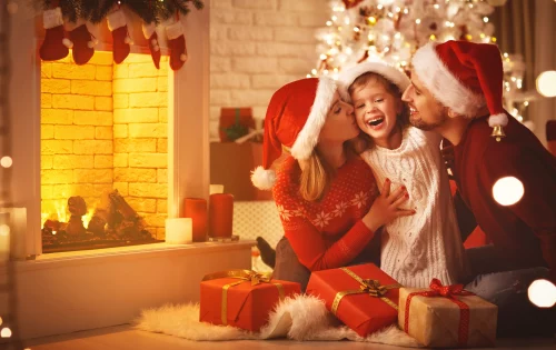 Imagem mostra um casal vestindo toucas de papai noel, abraçando uma criança feliz, em uma sala com lareira, árvore de natal, enfeites e pacotes de presente
