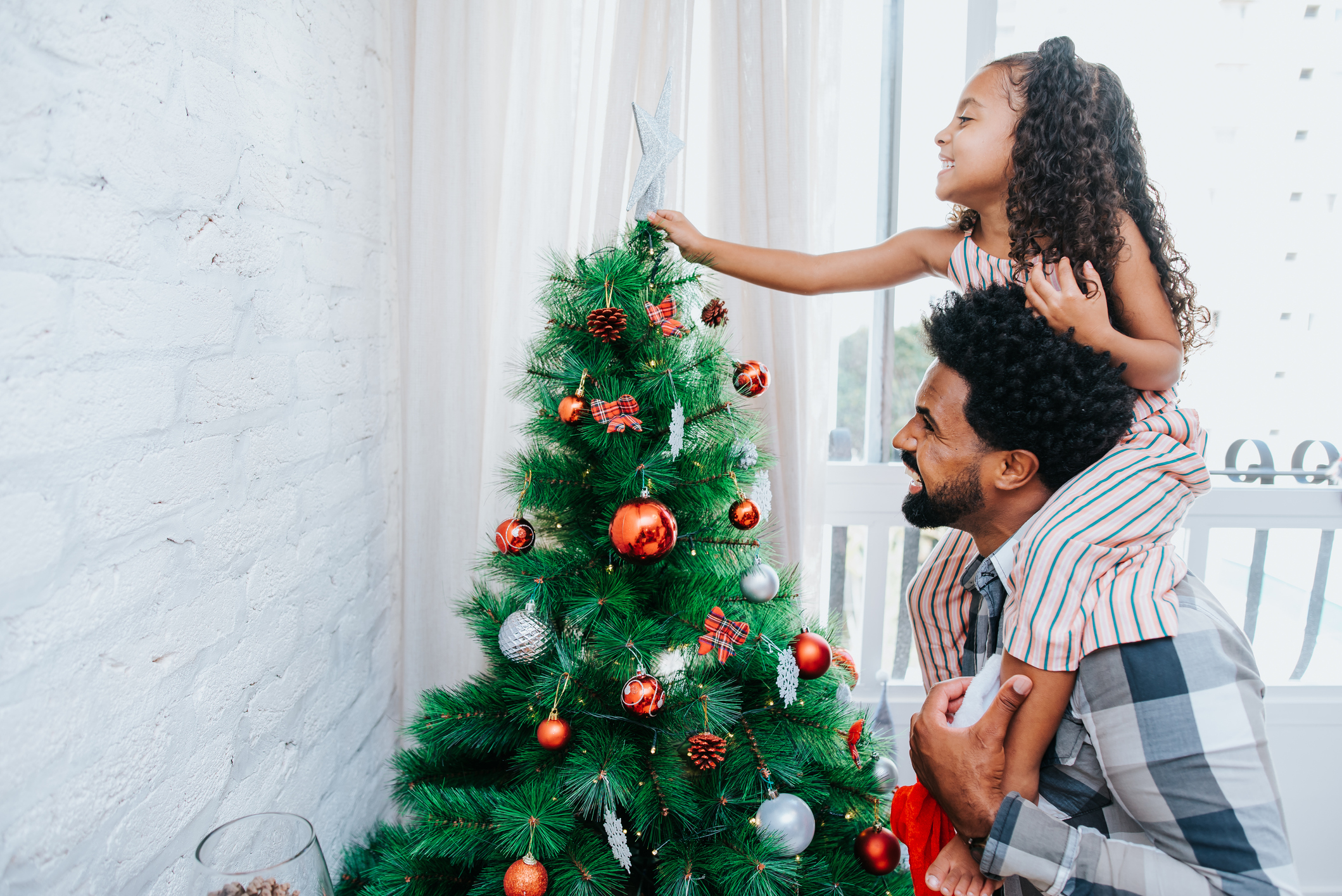 Imagem mostra uma criança nos ombros do pai colocando uma estrela no topo de uma árvore de natal decorada com bolas de diversas cores 